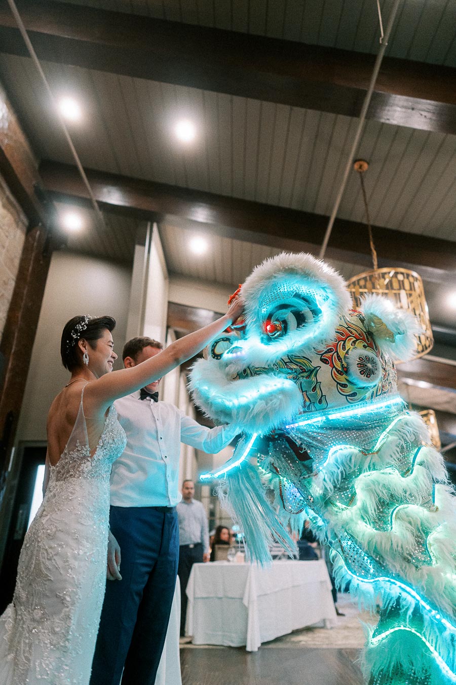 Bride and groom interacting with a vibrant, illuminated lion dance costume indoors during a wedding celebration.