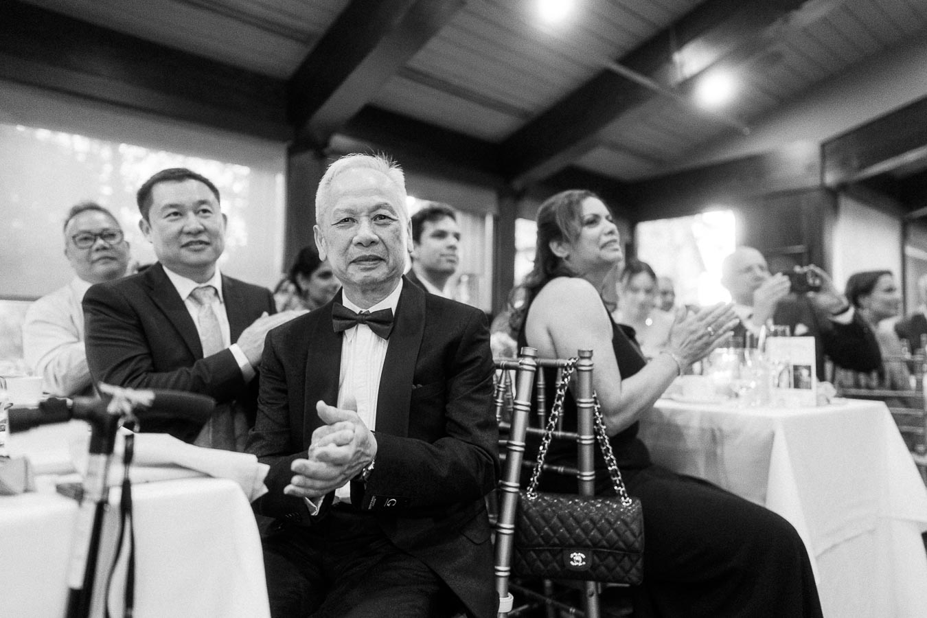 A group of elegantly dressed guests clapping during a formal event, seated at tables in a well-lit venue. The atmosphere is celebratory and upscale.