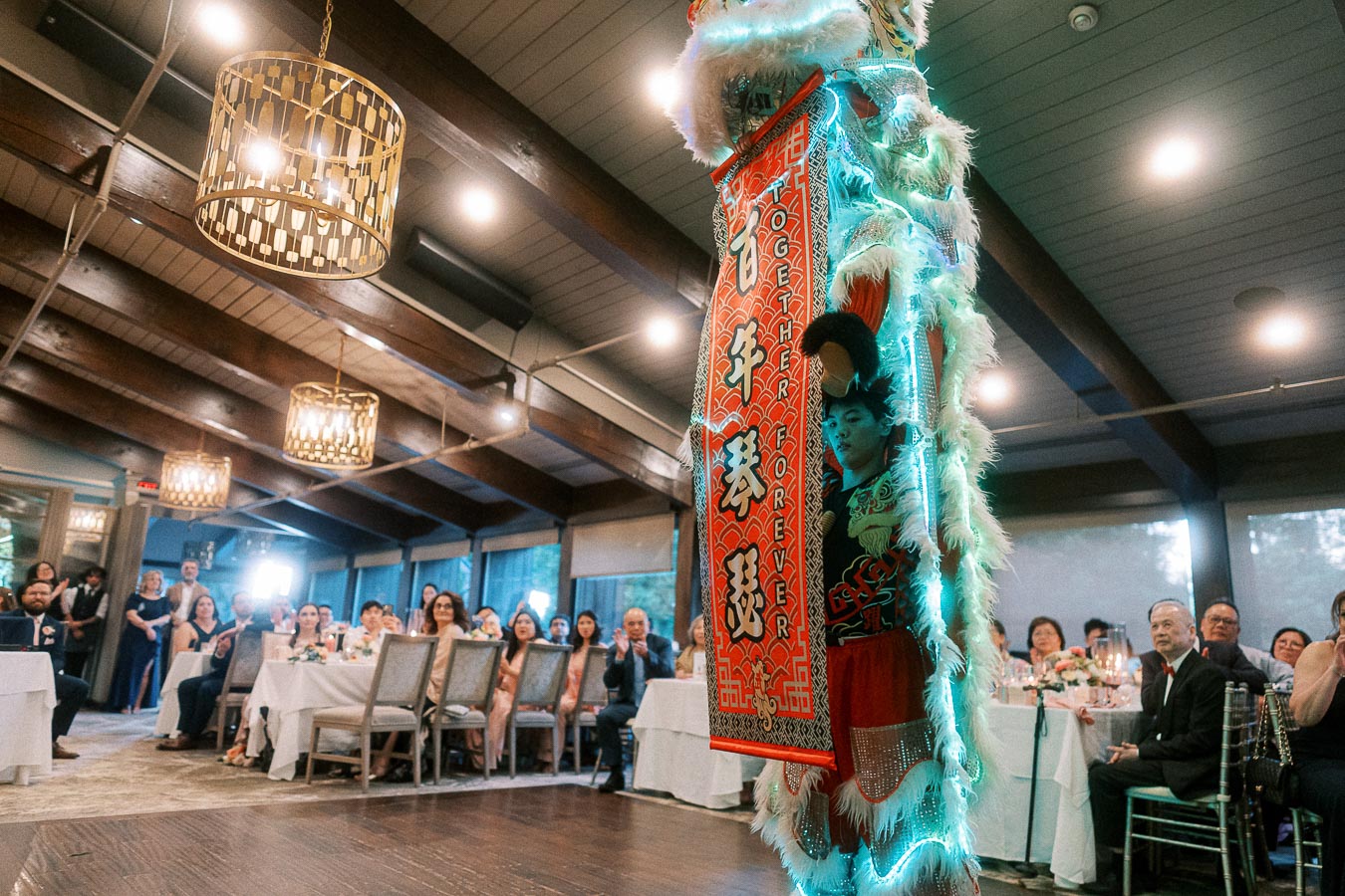 Lion dance performance at a wedding reception, featuring a vibrantly colored costume with illuminated details in a warmly lit venue. Guests, seated at decorated tables, watch attentively, enhancing the celebratory atmosphere.