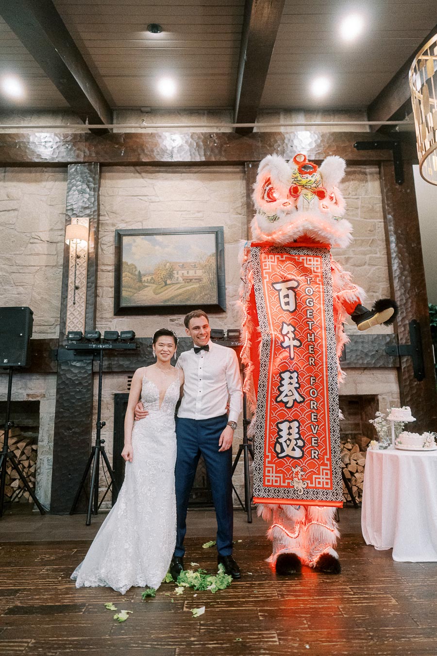 A bride and groom stand smiling next to a traditional Chinese lion dance performance at their wedding reception, with a festive banner reading Together Forever.