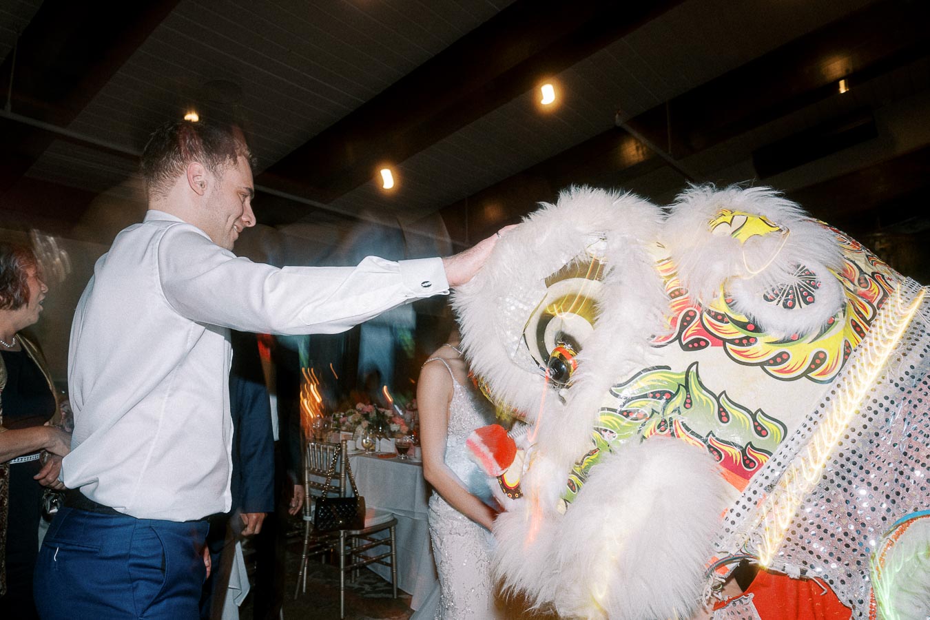 A person in a white shirt and blue pants interacts with a vibrant, ornate lion dance costume during a festive indoor event.