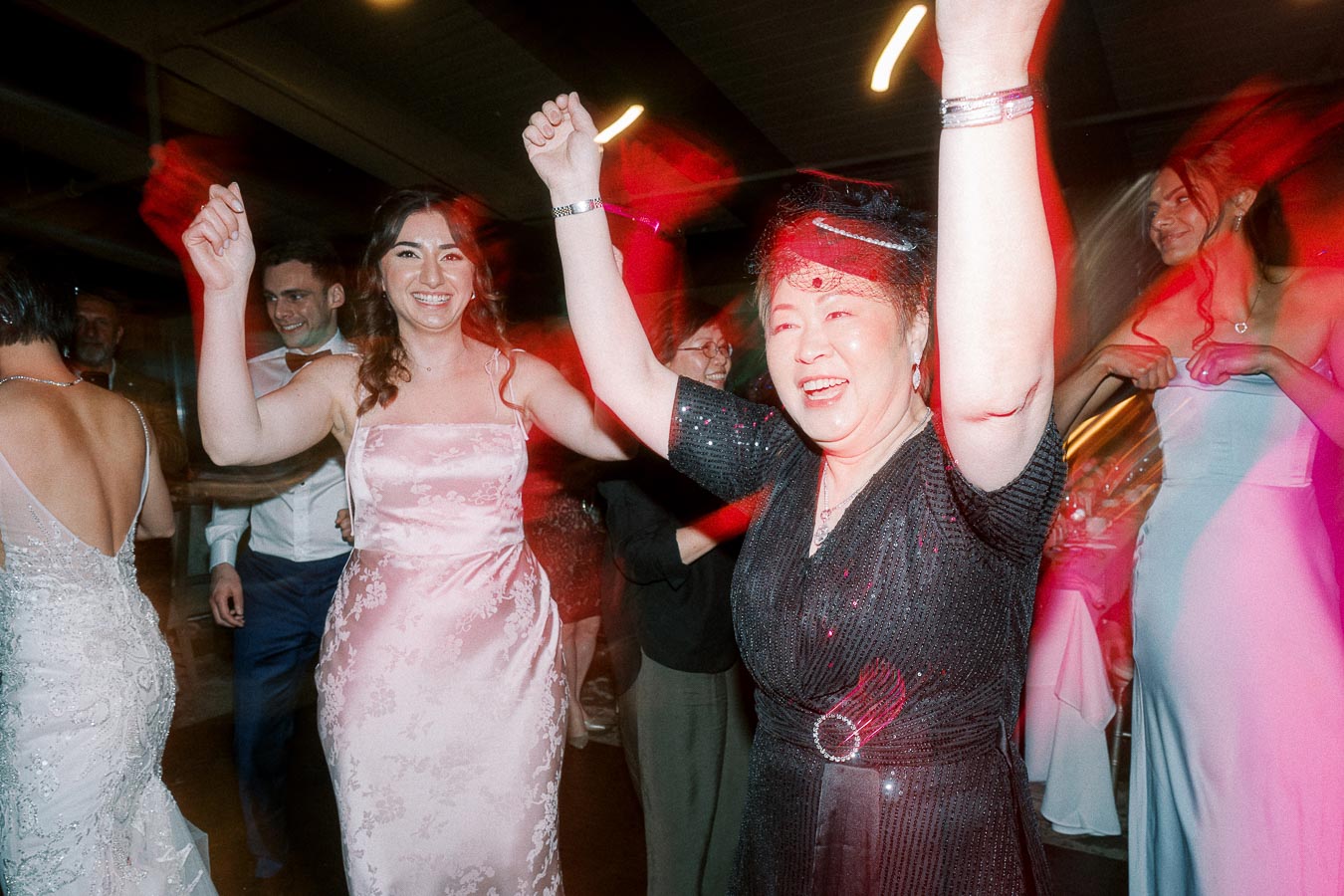 Joyful people dancing at a lively celebration, women in elegant dresses smiling and raising their arms in excitement, colorful lights enhancing the festive atmosphere.