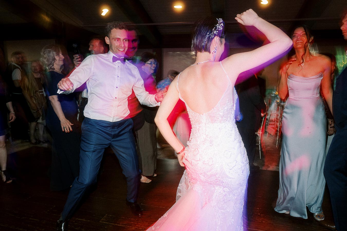 A lively wedding reception dance floor with a smiling groom in a white shirt and bow tie dancing with the bride in a stunning lace gown, surrounded by joyous guests in elegant attire.