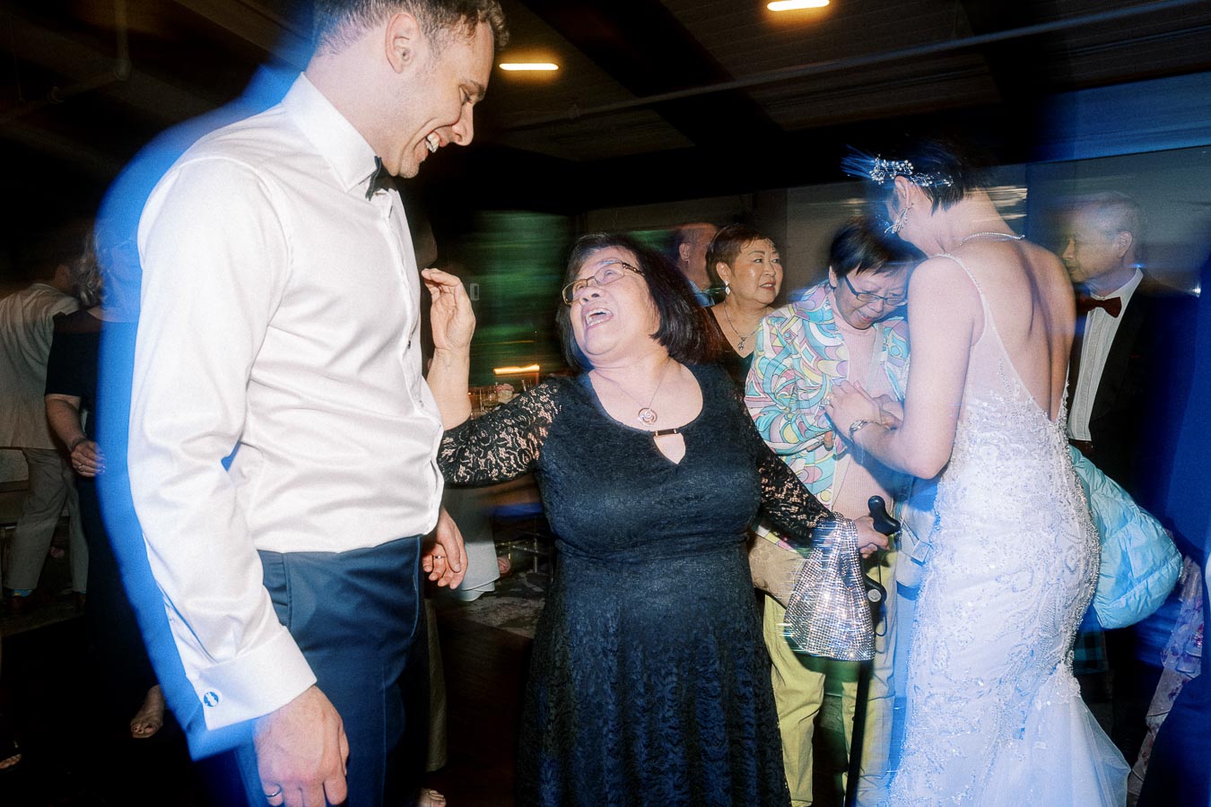 A joyful wedding celebration with guests dancing, featuring a woman in a black dress, a man in a white shirt, and a bride in a white gown enjoying the festive atmosphere.