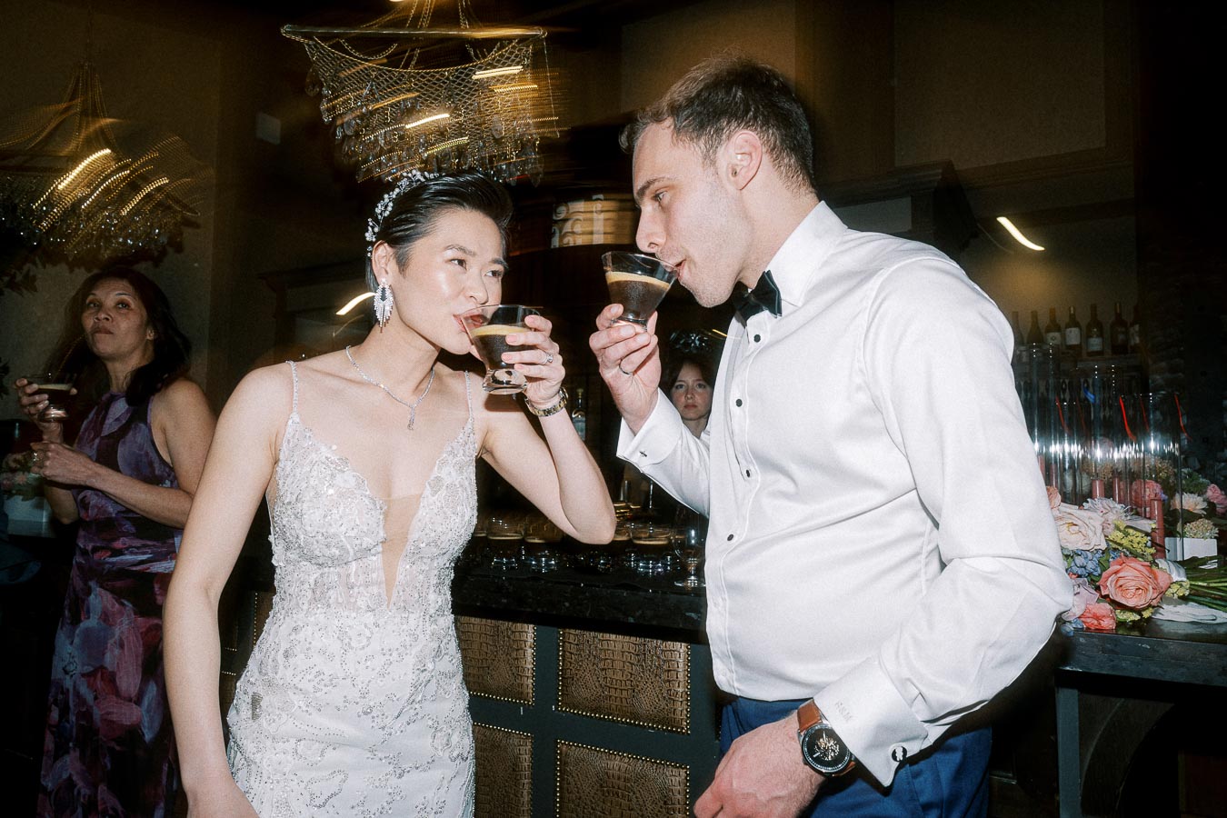 A bride and groom in elegant attire enjoying cocktails at their wedding reception, with a guest in the background and a decorative chandelier overhead.
