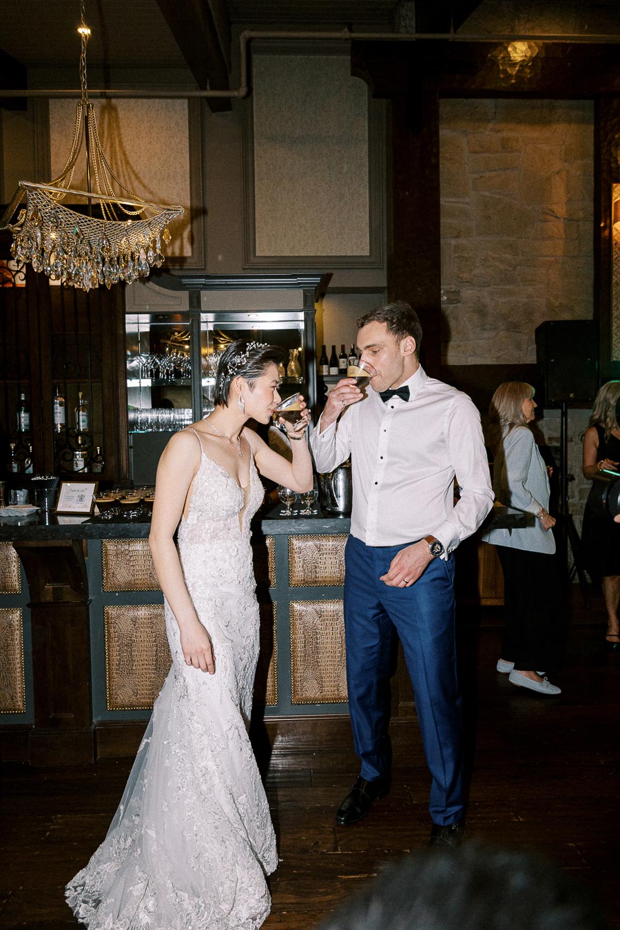 Bride and groom enjoying drinks together at an elegant reception with a chandelier and a bar in the background.