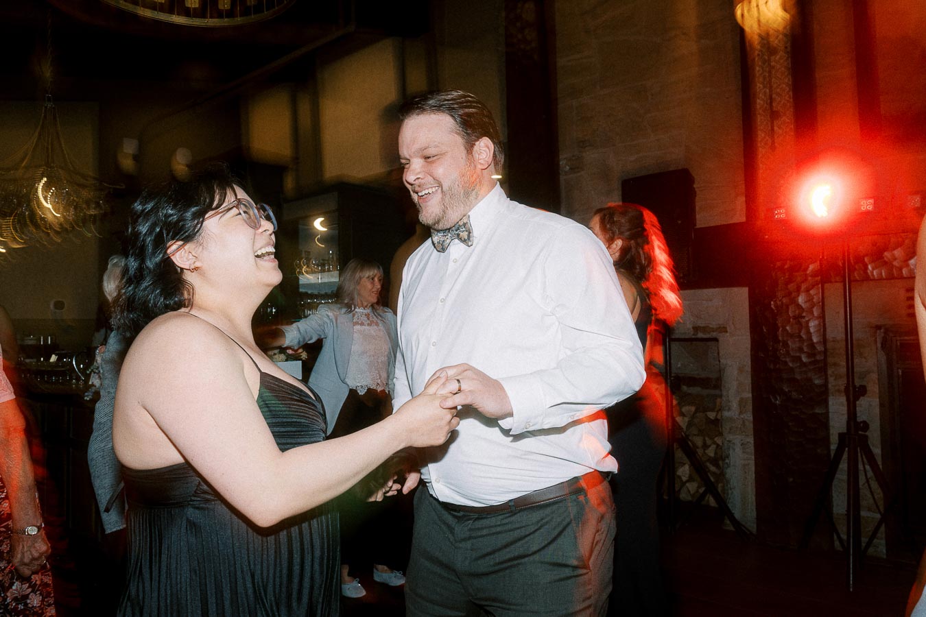 A joyful couple dancing and smiling at a reception, illuminated by warm lighting and surrounded by other guests enjoying the celebration.