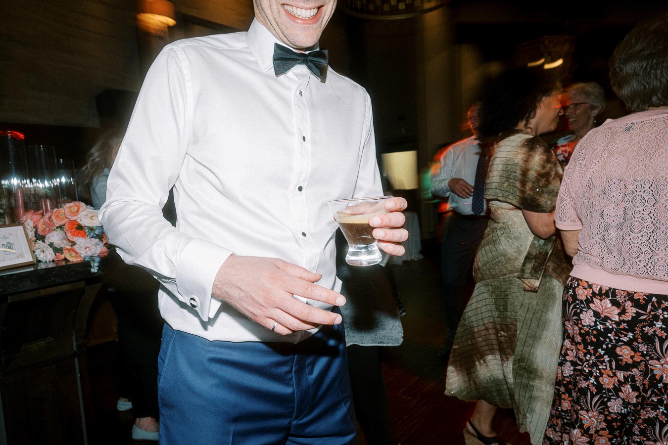 Man in a white dress shirt and bow tie holding a drink at a lively social event with people mingling and floral decorations in the background.