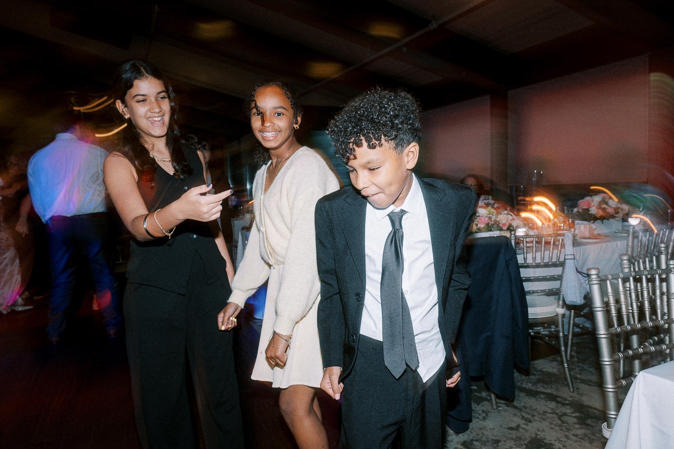 Three children smiling and dancing at a wedding reception, wearing formal attire with tables and chairs in the background.