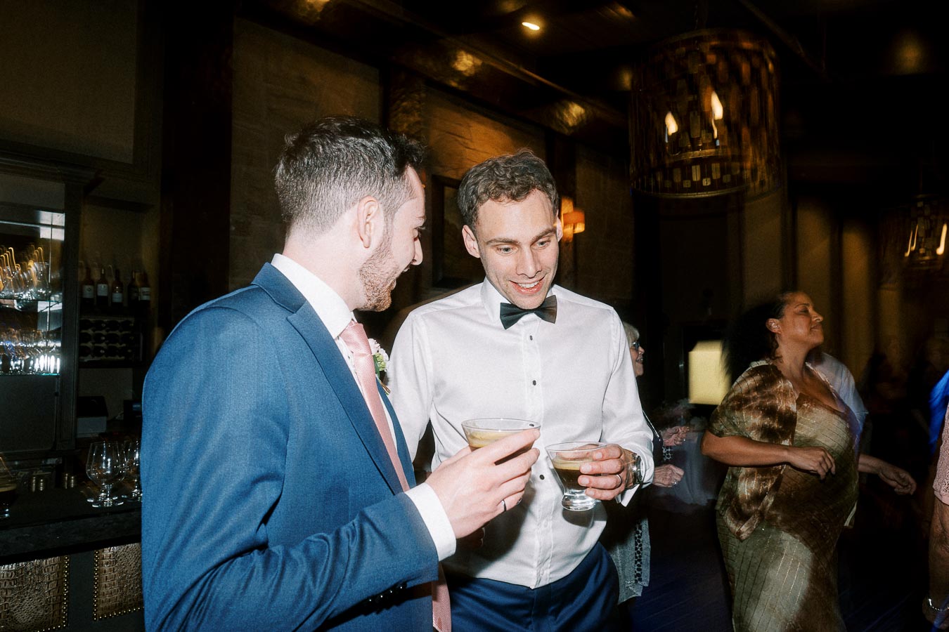 Two men in formal attire enjoy cocktails at a lively evening event, with ambient lighting and a festive atmosphere in the background.