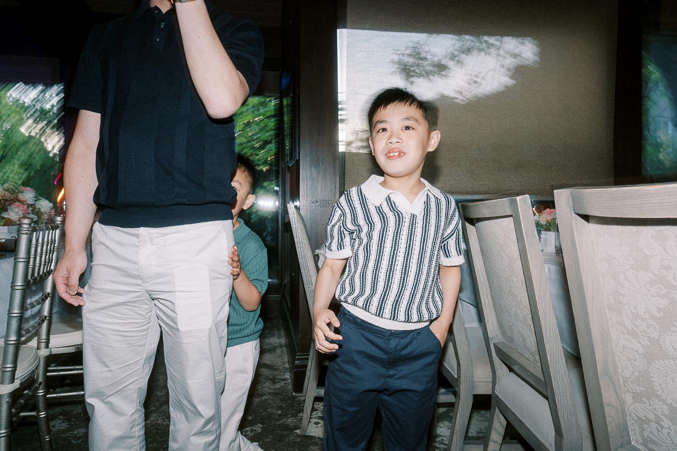 Young boy smiling at a family gathering while another child playfully hides behind an adult, inside a banquet hall with elegant seating arrangements.