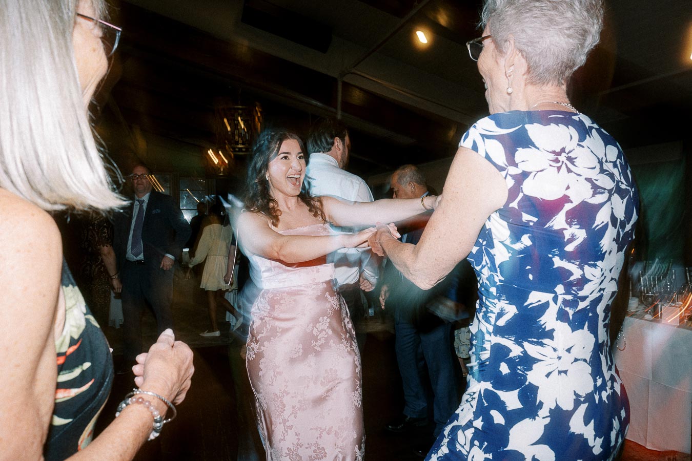 Lively wedding reception dance floor with guests enjoying a joyful dance, featuring a woman in a pink floral dress and another in a blue and white patterned dress, surrounded by a joyful crowd.