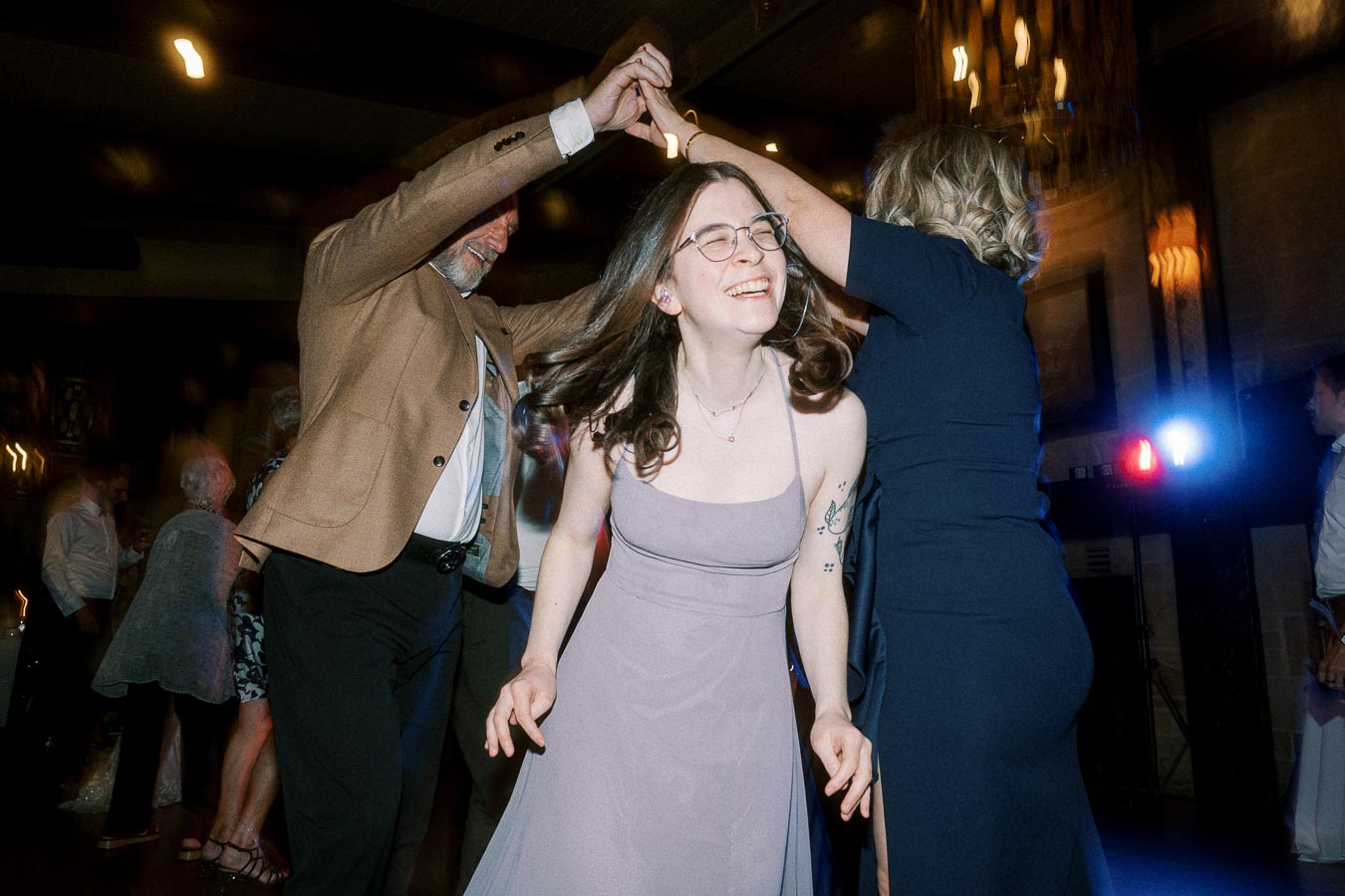 Group of people dancing joyfully at a lively indoor event, featuring a woman in a lavender dress smiling with closed eyes while others dance around her.