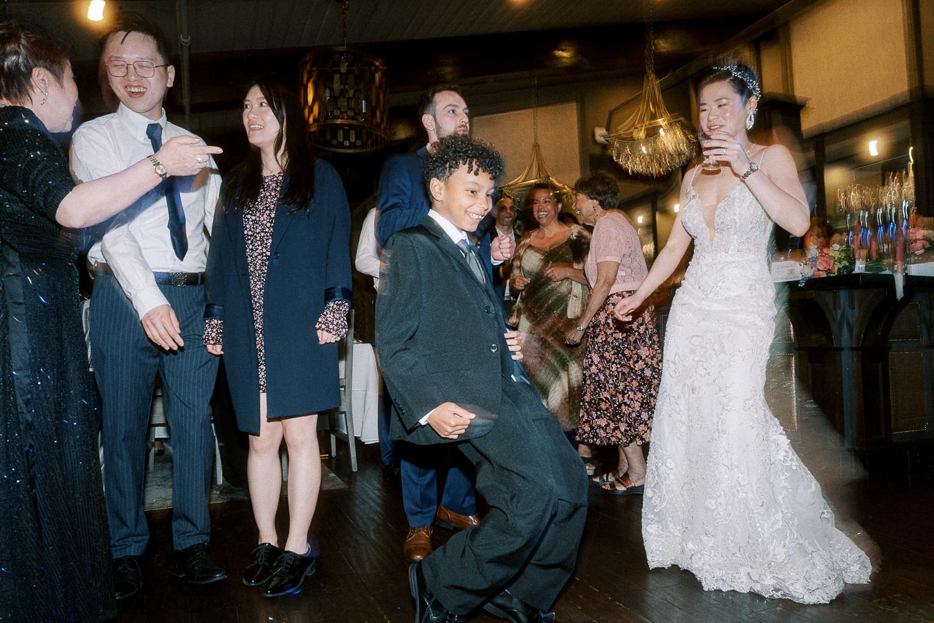 Group of people dancing and celebrating at a lively indoor wedding reception, with a smiling bride in an elegant white gown and guests in formal attire enjoying the festive atmosphere.