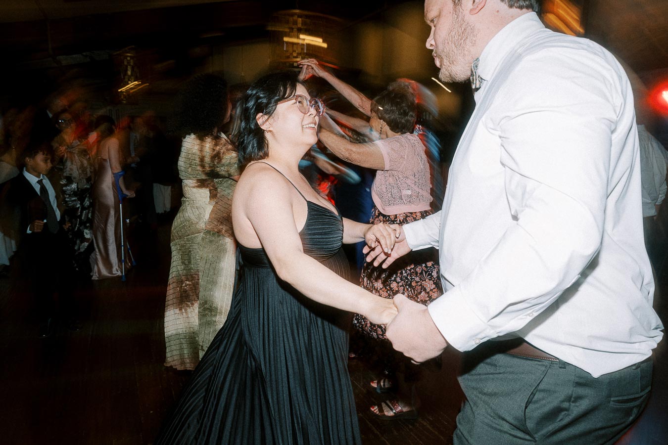 A joyful wedding reception dance floor with a couple in formal attire enjoying a dance, surrounded by other guests wearing elegant dresses and suits.