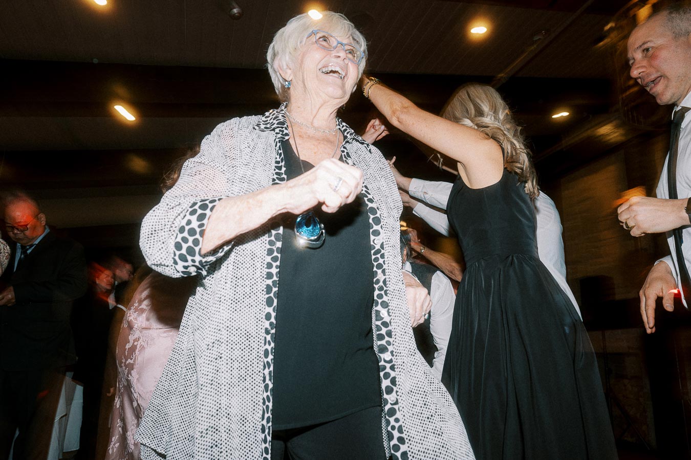 Elderly woman joyfully dancing at a lively indoor party with friends in formal attire.
