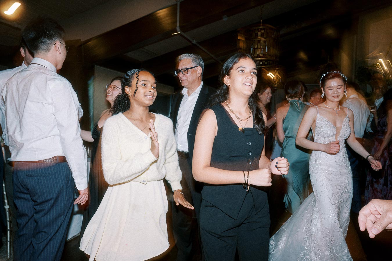 A lively wedding reception dance floor with diverse guests having fun, including a smiling woman in a white dress, another in a black outfit, and a bride in an elegant white gown, all enjoying the celebration.