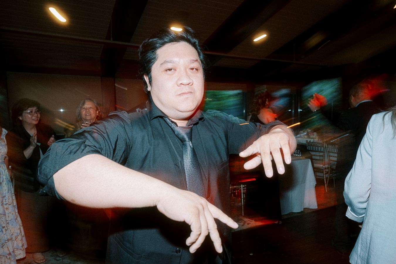 A person in a black shirt and tie dancing energetically at a lively indoor party, with other guests in the background smiling and clapping. The room is adorned with ambient lighting and tables set up for the event.