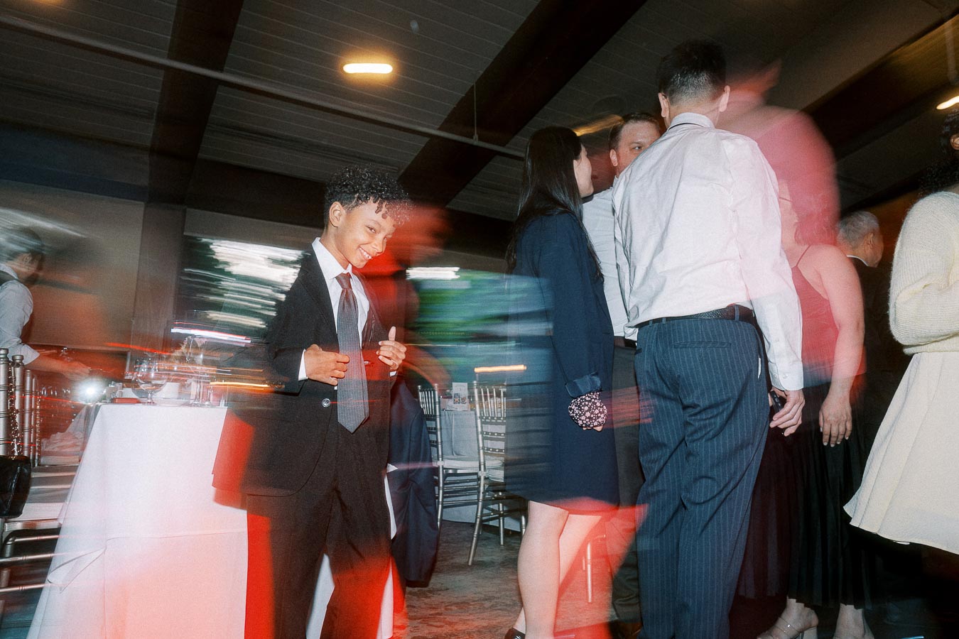 A young boy in a suit dancing joyfully at an indoor event, surrounded by a blurred group of elegantly dressed people, under ambient lighting.