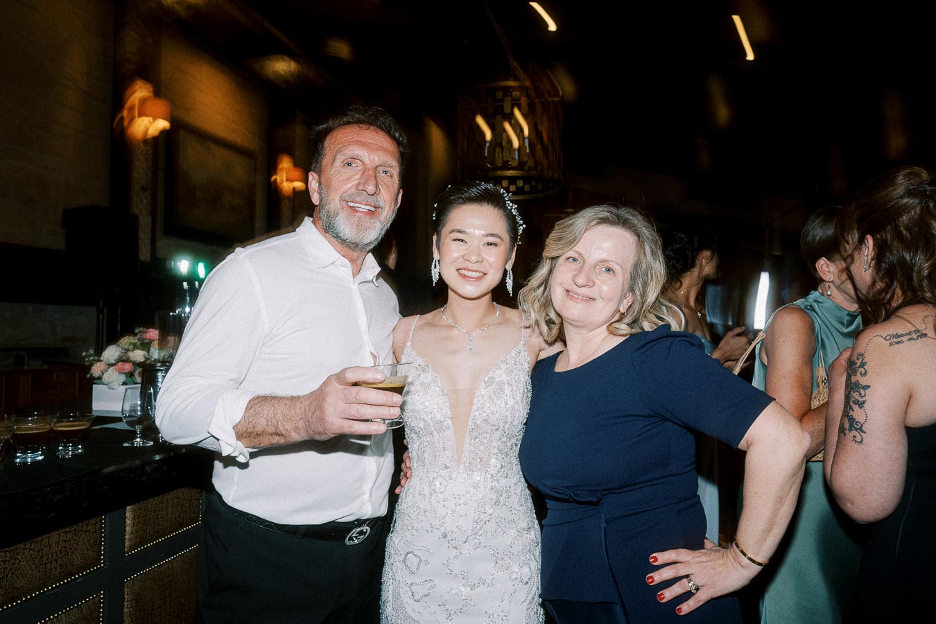 A smiling bride in a white dress poses with two guests at a wedding reception.
