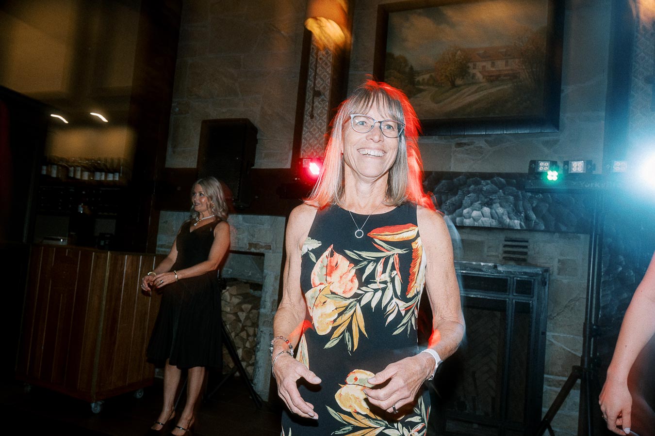 A woman smiling and wearing a floral dress, attending a party indoors with ambient lighting and a rustic fireplace in the background.