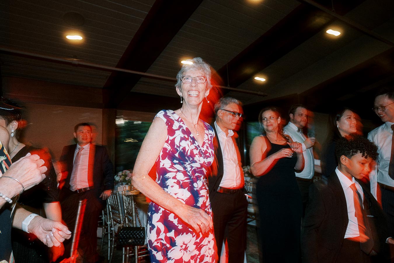Senior woman smiling in floral dress amidst group of people at evening indoor event.