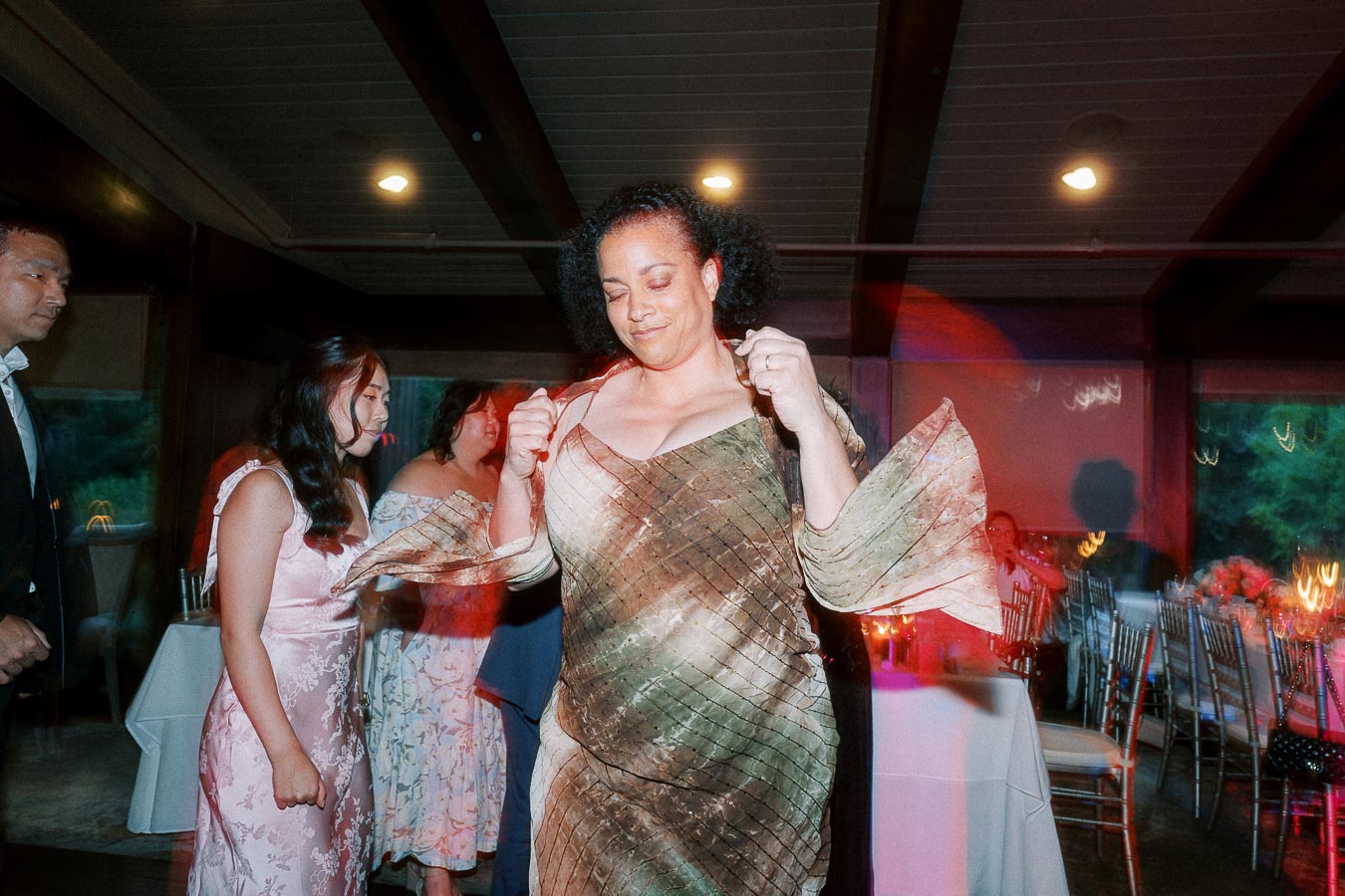Woman in elegant dress dancing joyfully at a wedding reception, surrounded by guests in a warmly lit event space.