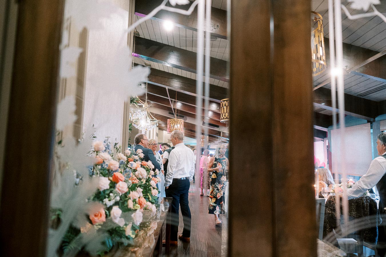 Elegant event gathering in a banquet hall with guests socializing, featuring floral arrangements and warm ambient lighting.