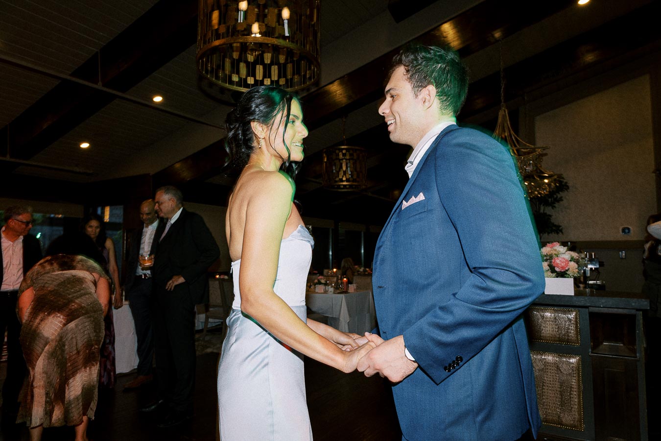 A couple elegantly dressed, holding hands and smiling while dancing at a lively indoor event, surrounded by guests in formal attire and under warm, ambient lighting.