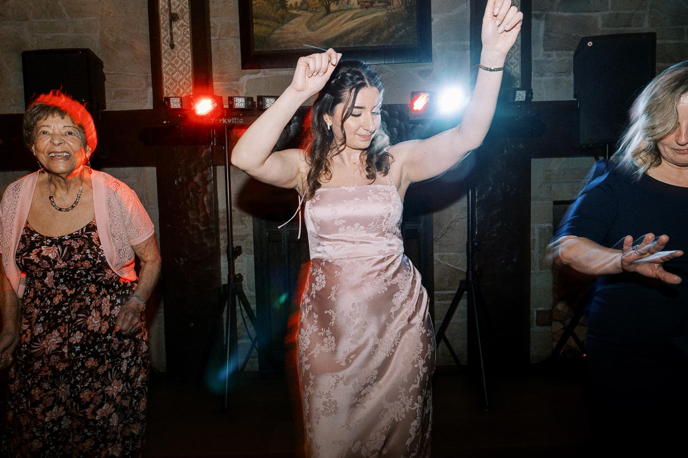 Three women joyfully dancing at a lively indoor party, dressed in elegant outfits, with colorful lights in the background.