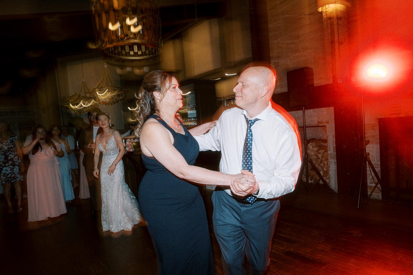 A couple dancing at a wedding reception, surrounded by guests in elegant attire, with warm lighting creating a festive atmosphere.
