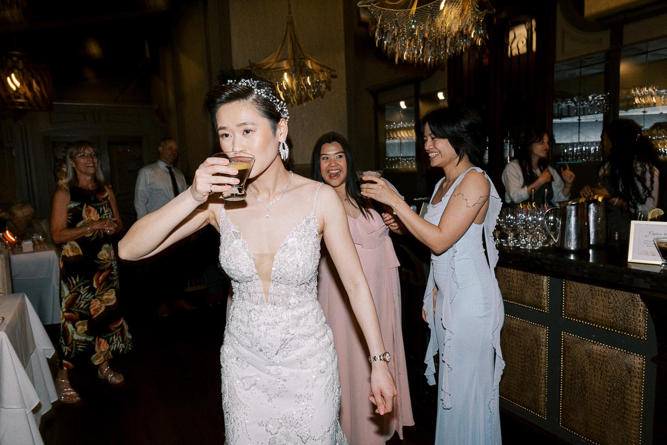 A bride in a detailed lace wedding gown enjoys a drink at a lively wedding reception, with guests smiling and celebrating in the background near a well-stocked bar.