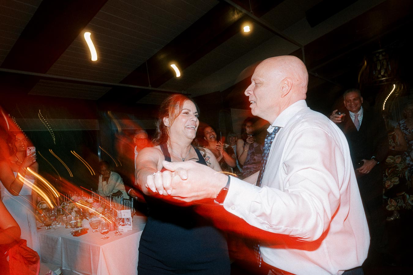 A joyful couple dancing energetically at a lively wedding reception, surrounded by smiling guests, with a beautifully decorated dining table visible in the background.