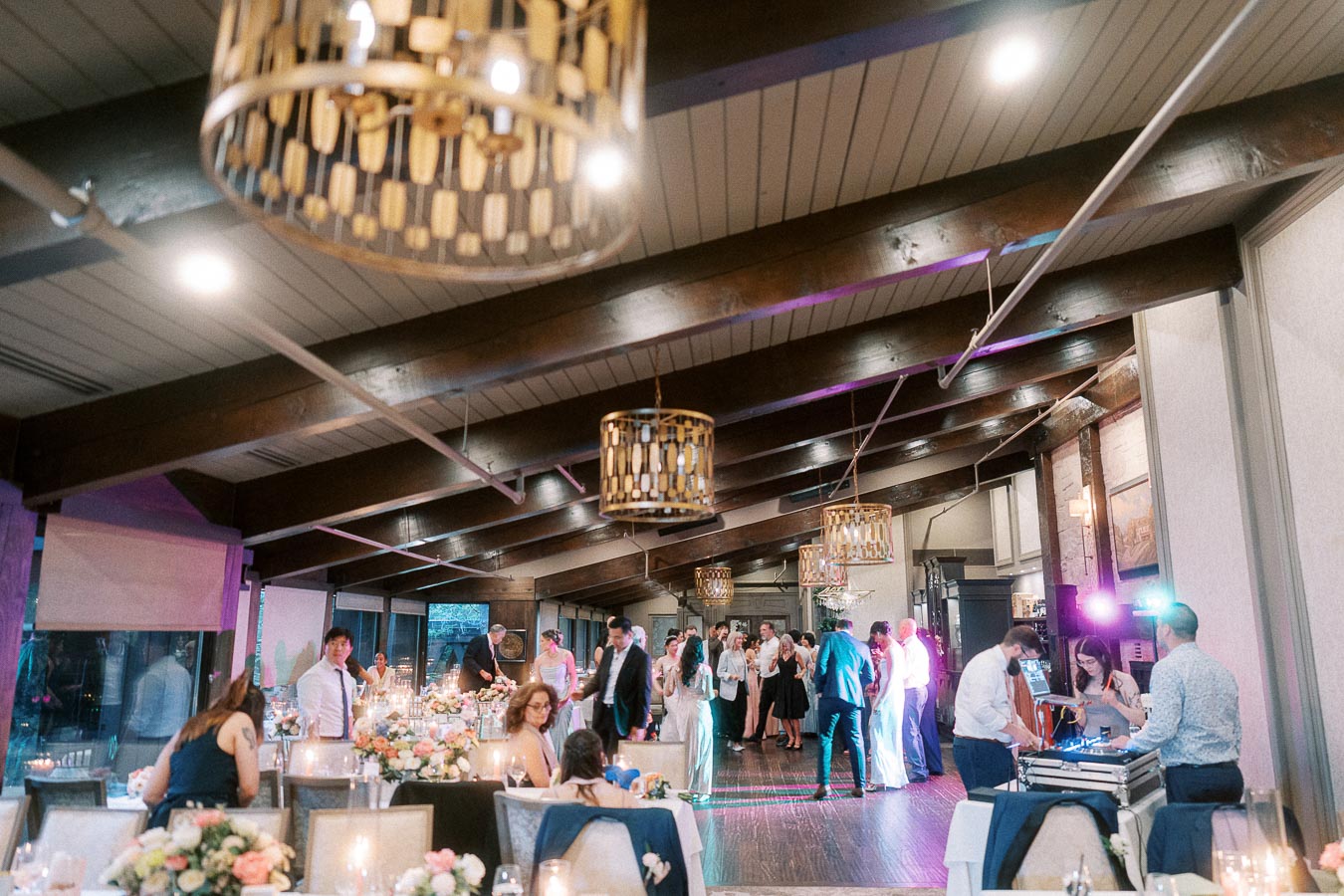 A lively wedding reception in a stylish venue with guests dancing and socializing under elegant lighting, surrounded by beautifully decorated tables with floral centerpieces.