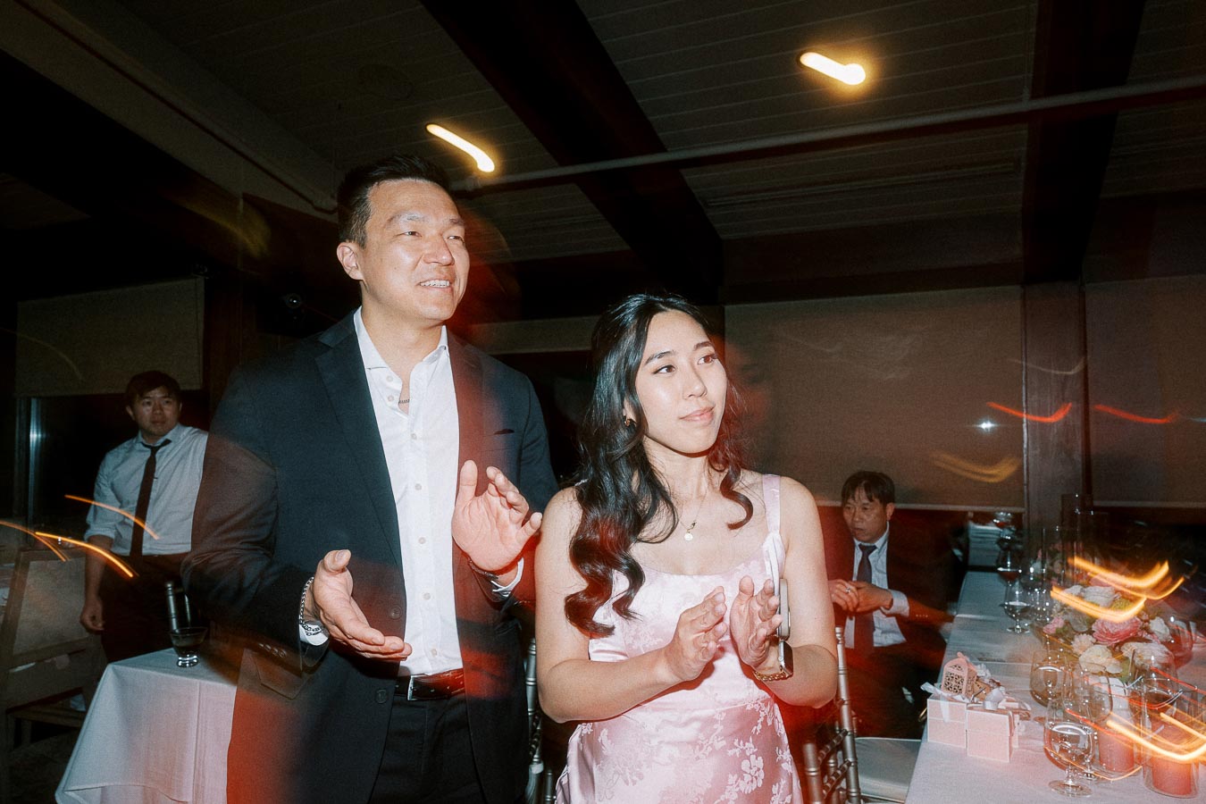 A couple in formal attire clapping at an indoor event, surrounded by guests and a decorated table.