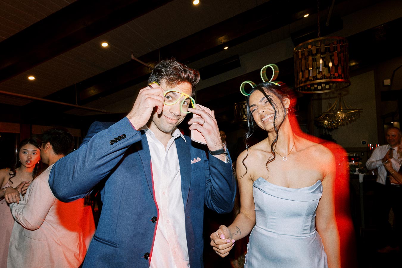 People enjoying a lively party with neon accessories, man in a blue suit playfully adjusts glowing glasses, while a woman in a silver dress smiles under a party atmosphere with colorful lighting and background dancers.