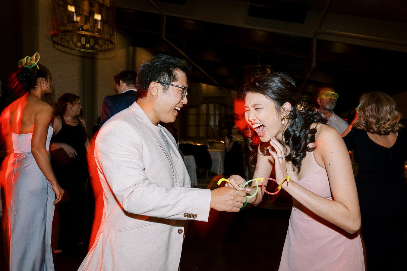 Guests laughing and dancing at a lively wedding reception, featuring cheerful interactions and party lights in a warmly lit venue.