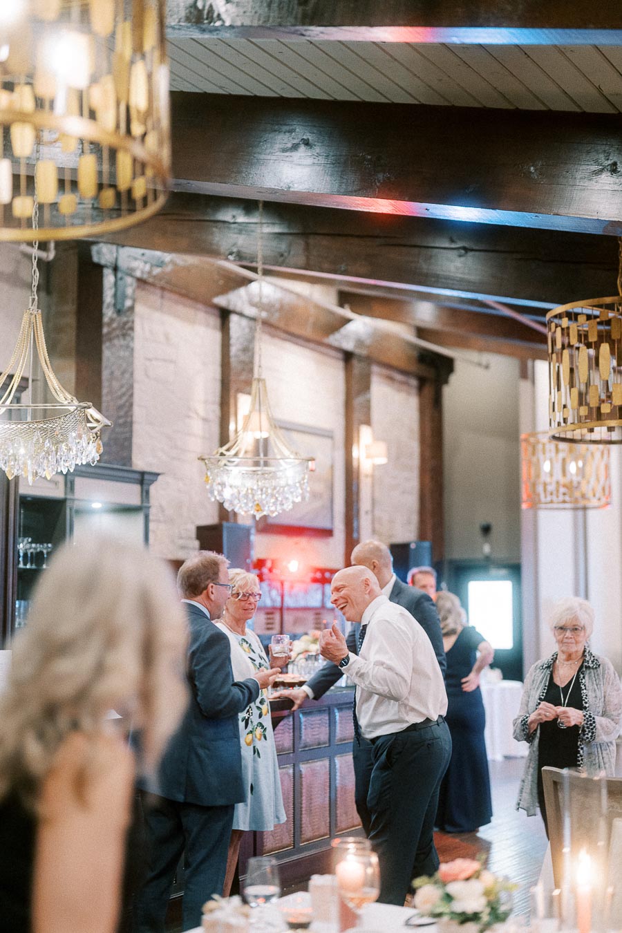 Elegant event scene with guests socializing in a warmly lit venue featuring chandeliers and rustic decor.