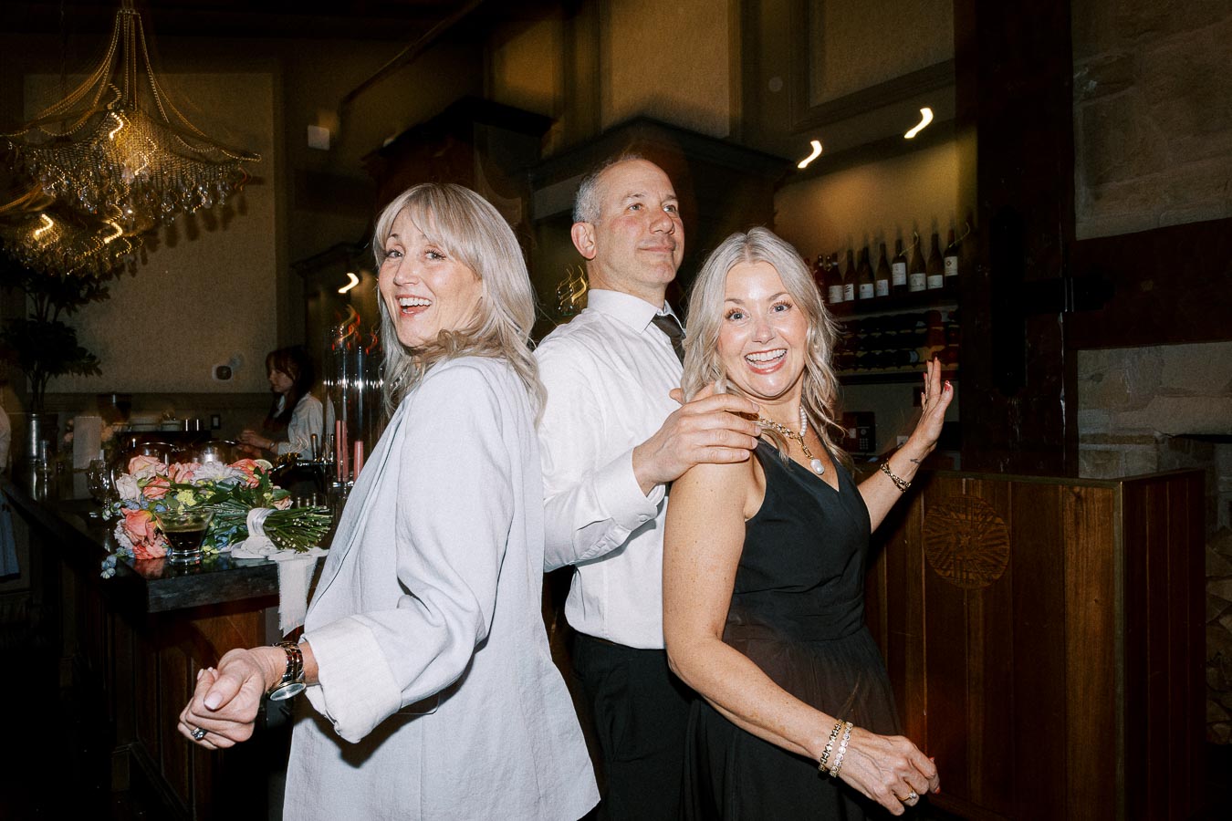 Three smiling people enjoying a festive gathering at a stylish restaurant, with elegant lighting and a beautifully set bar in the background.
