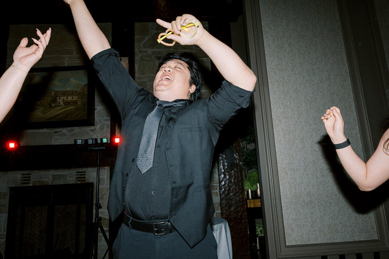 A person in a black shirt and tie, joyfully dancing at a lively indoor event, with arms raised and a big smile, capturing the festive ambiance of the celebration.