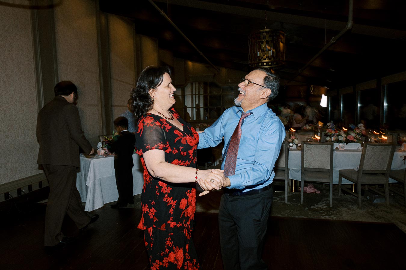 A joyful couple dancing together at an indoor event, with decorated tables and guests in the background.