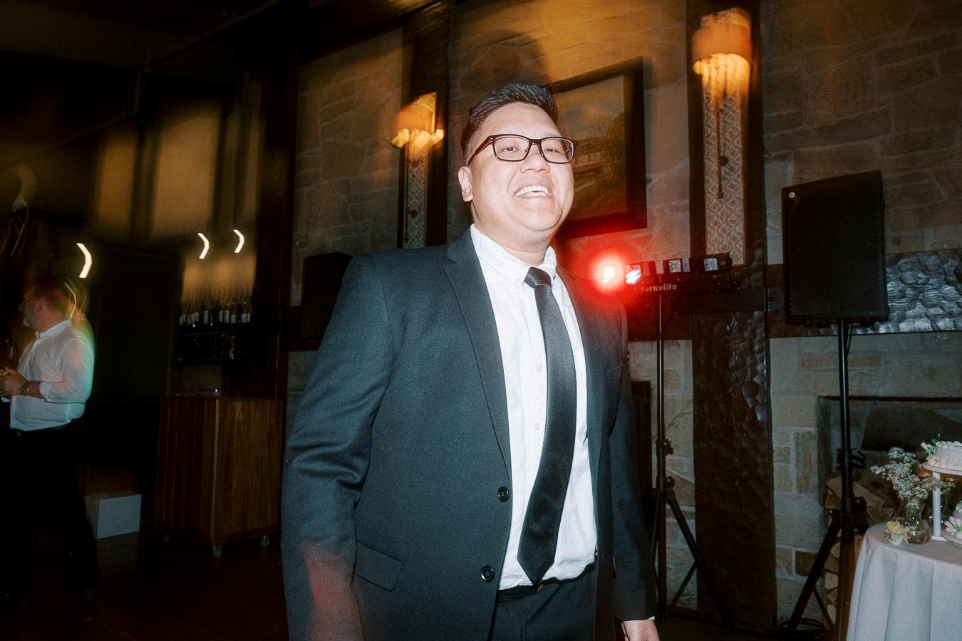 Man in a suit and tie smiling at an event in a warmly lit room, with stone walls and ambient lighting in the background.