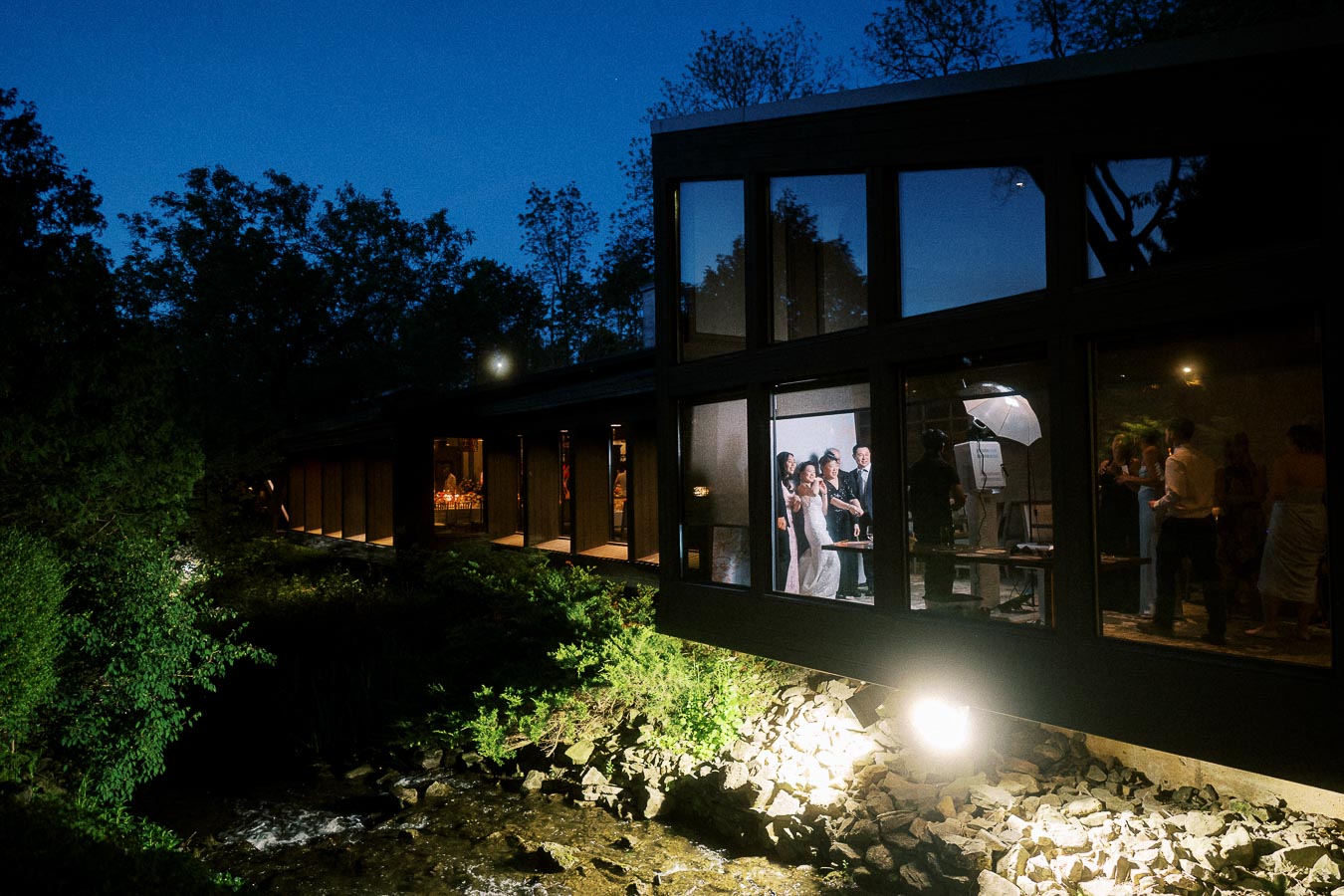 Exterior view of a modern building at night with large windows showing an indoor wedding reception. Guests are visible celebrating inside, surrounded by lush greenery and a gently flowing creek beneath.