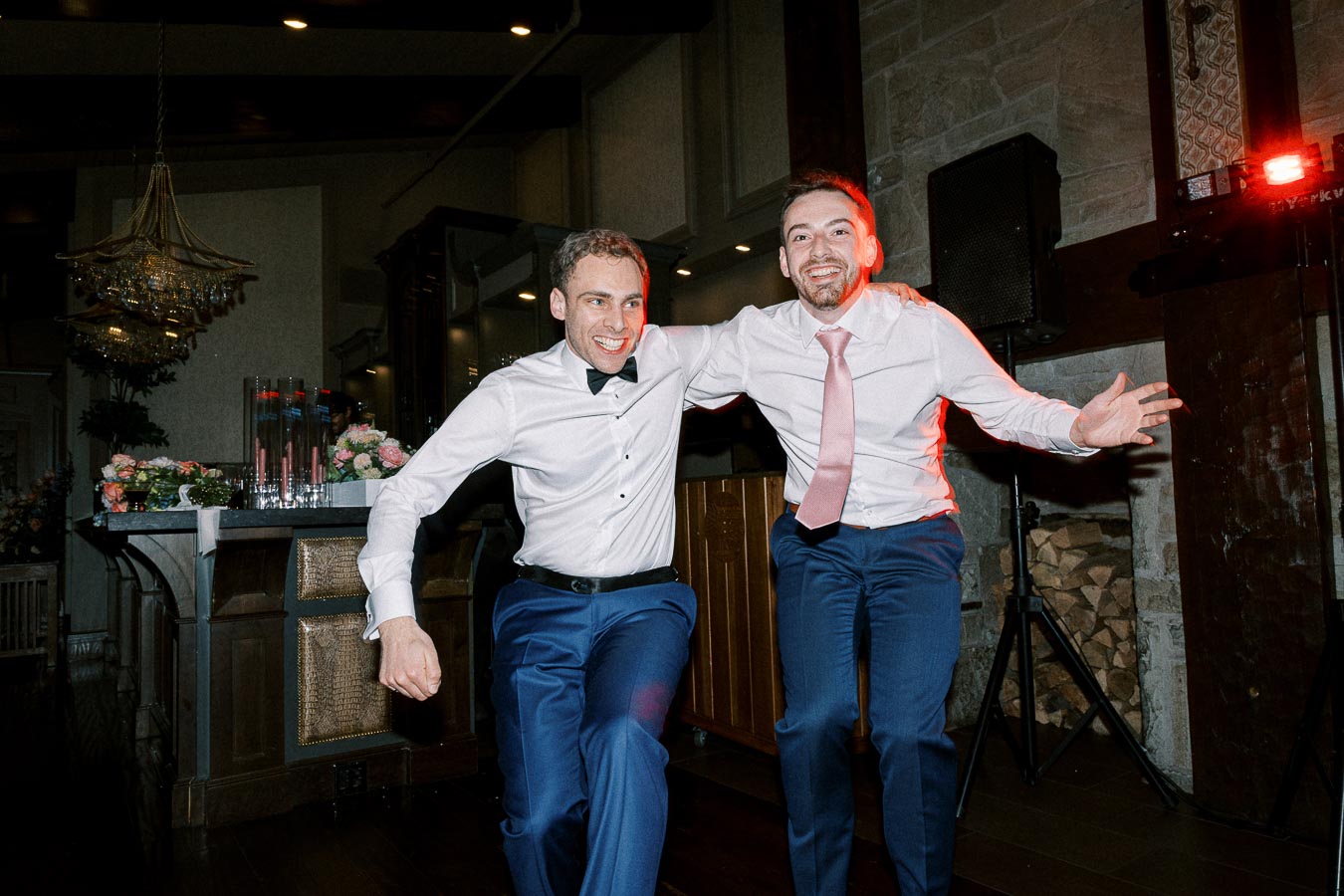 Two men in formal attire joyfully dancing at a lively indoor wedding reception.