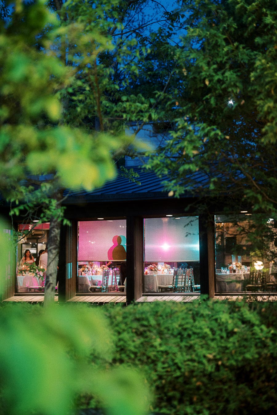 Exterior view of a beautifully lit restaurant with large windows, showcasing elegantly arranged tables inside. Lush greenery surrounds the building, creating a serene ambiance under the evening sky.