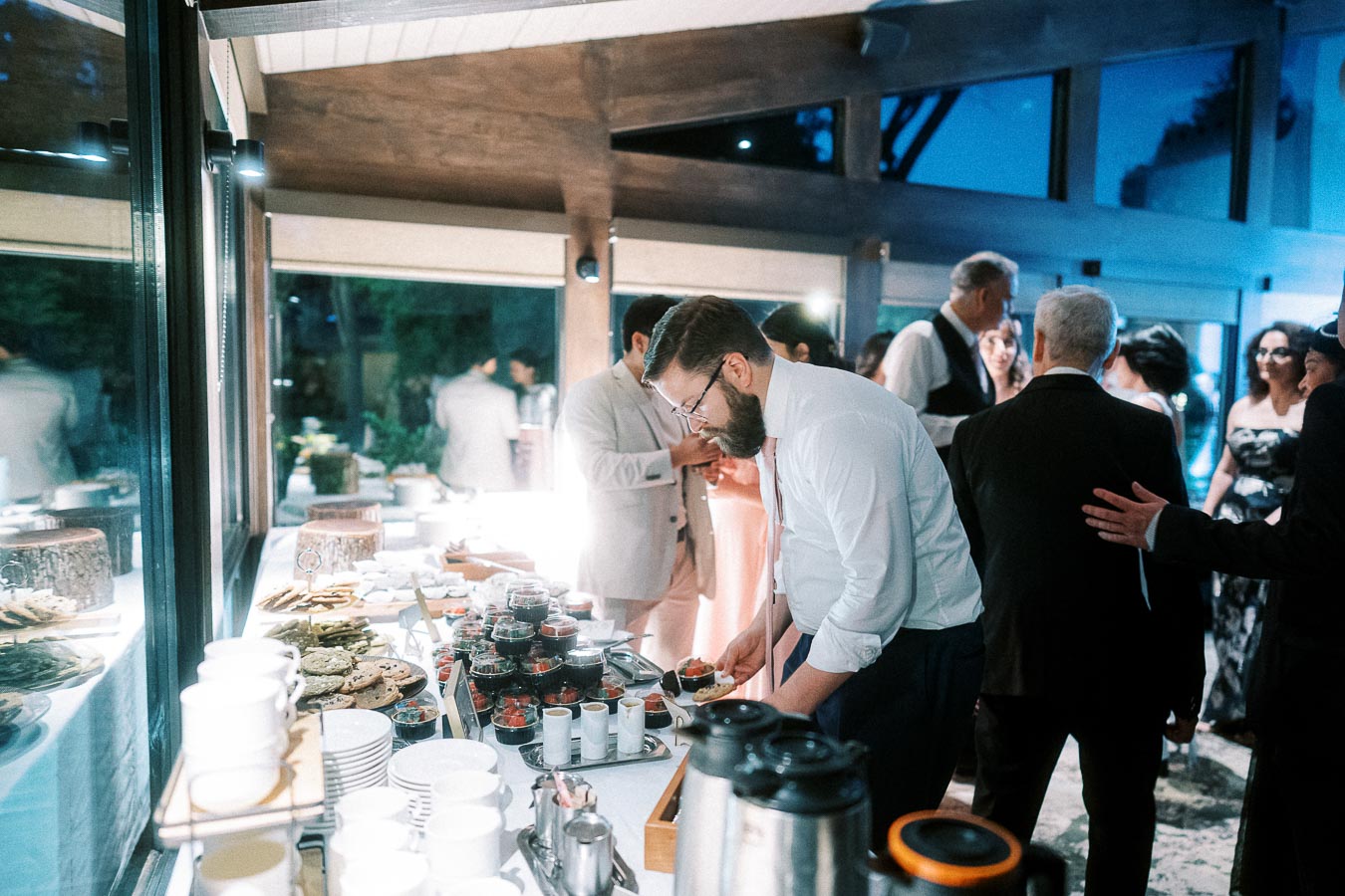 A group of elegantly dressed people gathers around a buffet table at a sophisticated indoor event. A man in a white shirt and tie is serving himself from an assortment of dishes, while others mingle and converse in the background. The atmosphere is warm and inviting, with a focus on the modern decor and the variety of food offerings.
