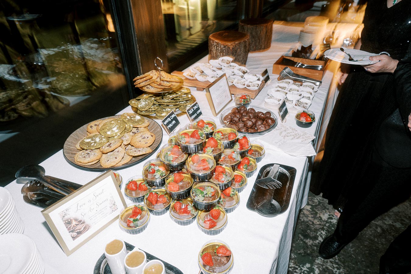Dessert table with a variety of sweet treats including cookies, chocolate-covered desserts, and fresh strawberries in cups. Plates and utensils are available for guests.