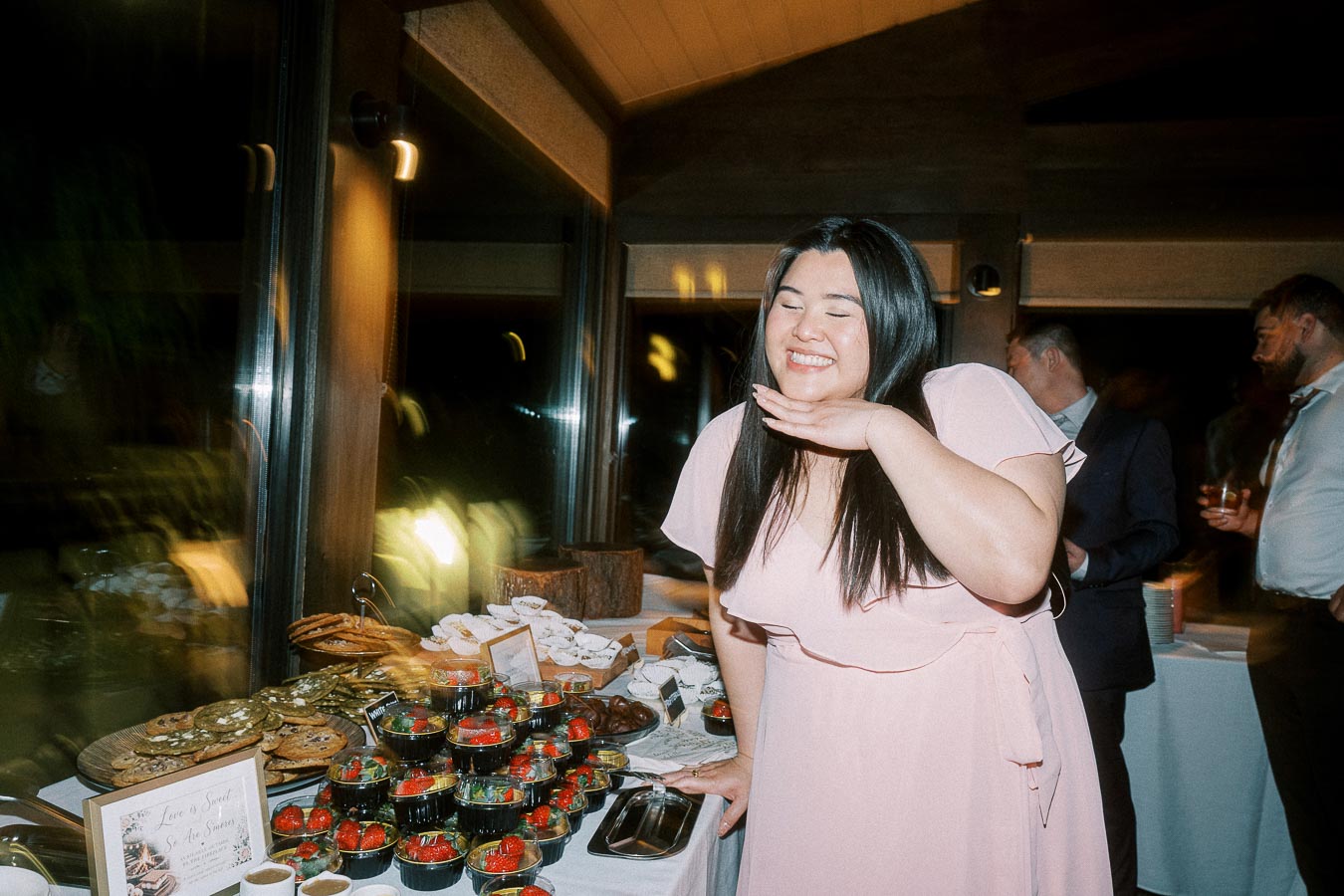A joyful woman in a pink dress posing with a delightful dessert spread, featuring cookies, strawberries, and assorted treats, at a festive event.