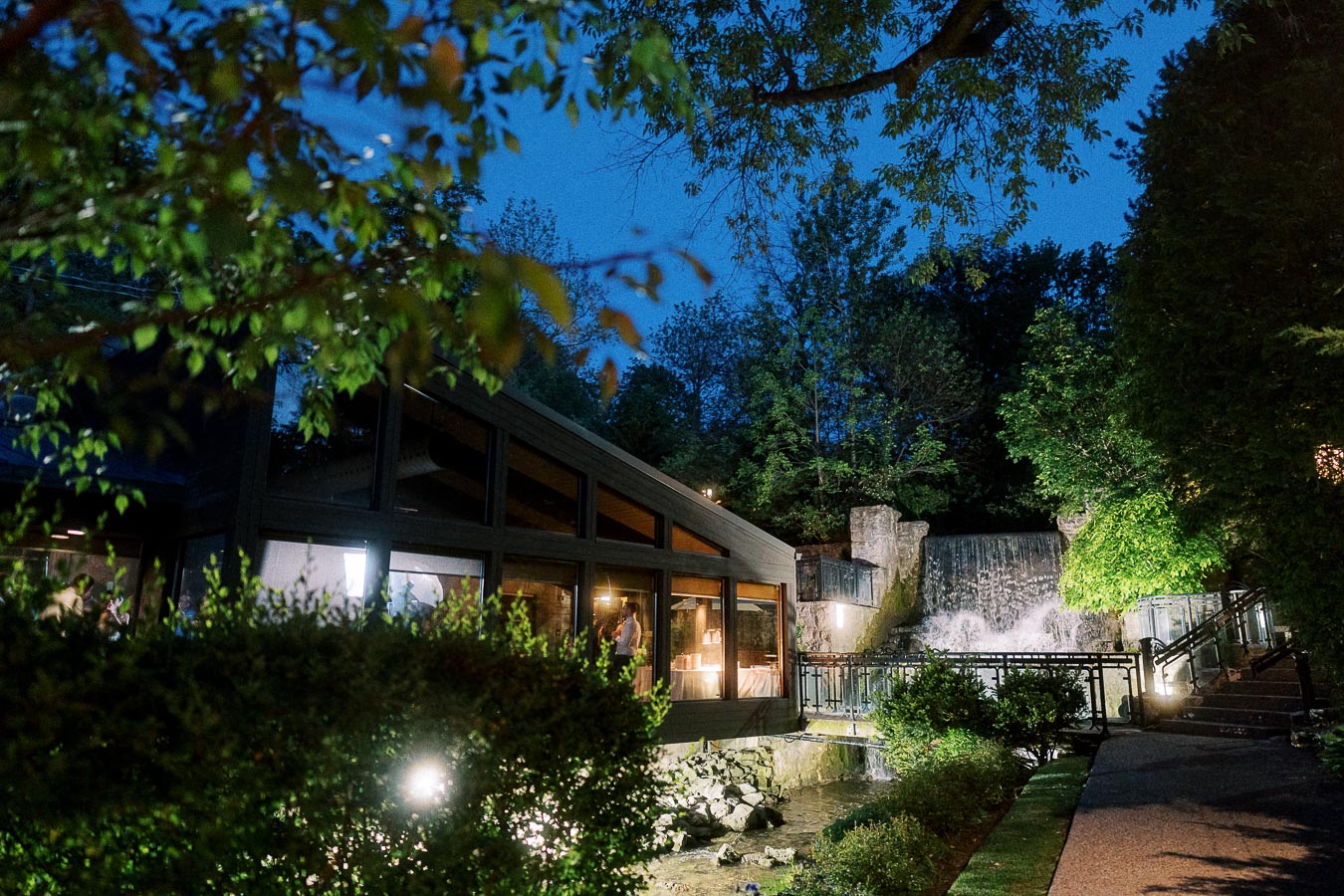 Modern restaurant with large windows illuminated at night, surrounded by lush trees and a softly lit waterfall in the background, creating a serene, picturesque outdoor setting.