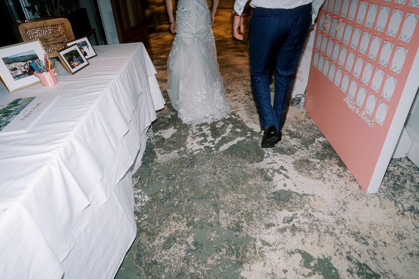 Bride and groom walking away from a decorated table at a wedding reception, featuring a pink seating chart on the right and framed photos on a white cloth-covered table to the left.